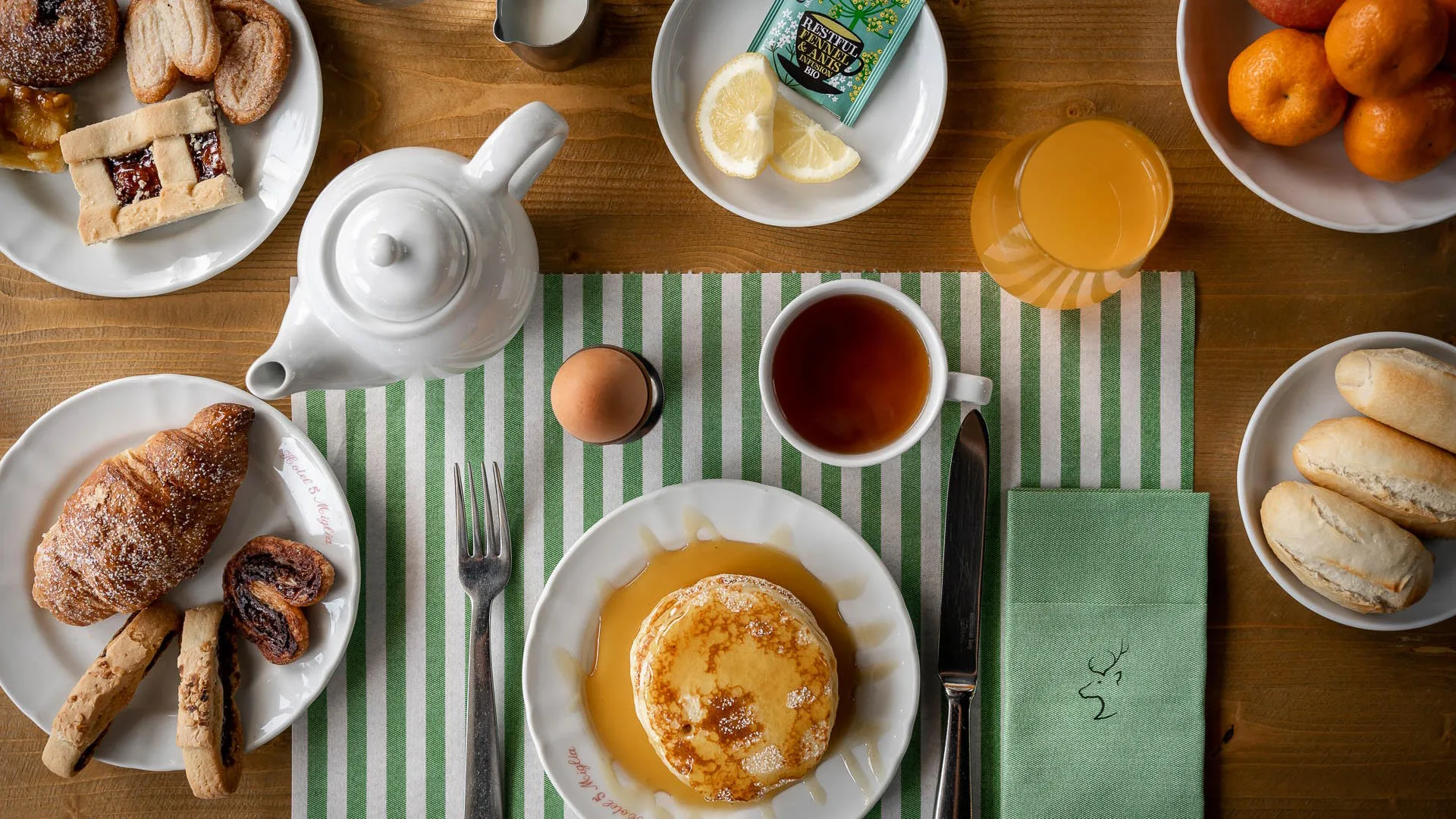 Tavolo apparecchiato per la colazione con pancake allo sciroppo, cornetto, tè, uovo, succo e frutta