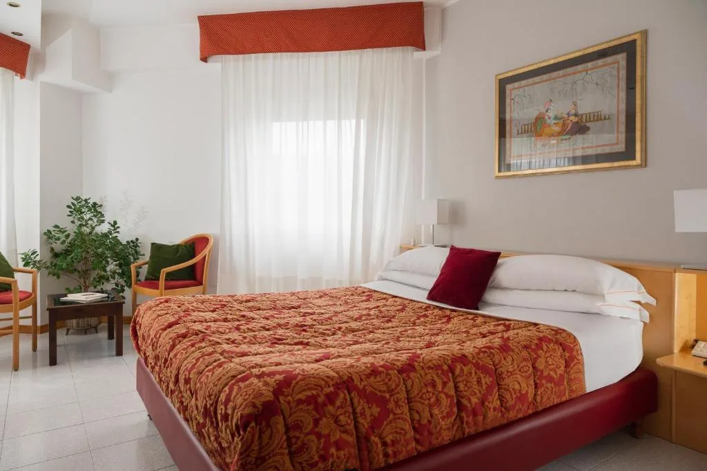 Camera matrimoniale con copriletto rosso e oro, finestra con tende bianche e poltrone