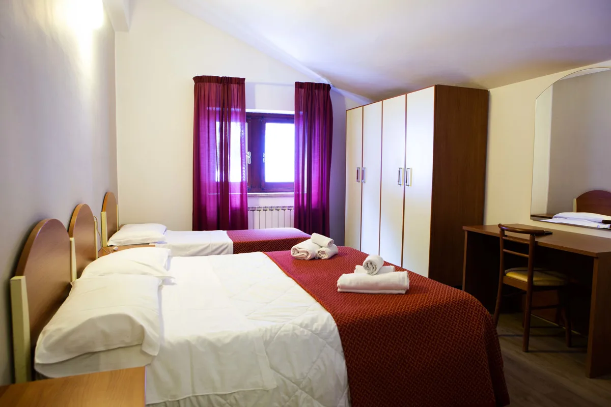 Camera con letti singoli, coperte rosse, armadio in legno e finestra con tende viola